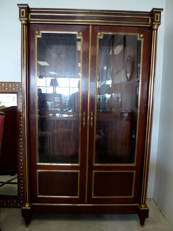 Majestic 19th century Louis XVI style biblioteque or bookcase in mahogany with gilt brass moldings and details. Two glass doors open to reveal optional glass shelves. <br />
<br />
A classic display piece for your leather bound books and