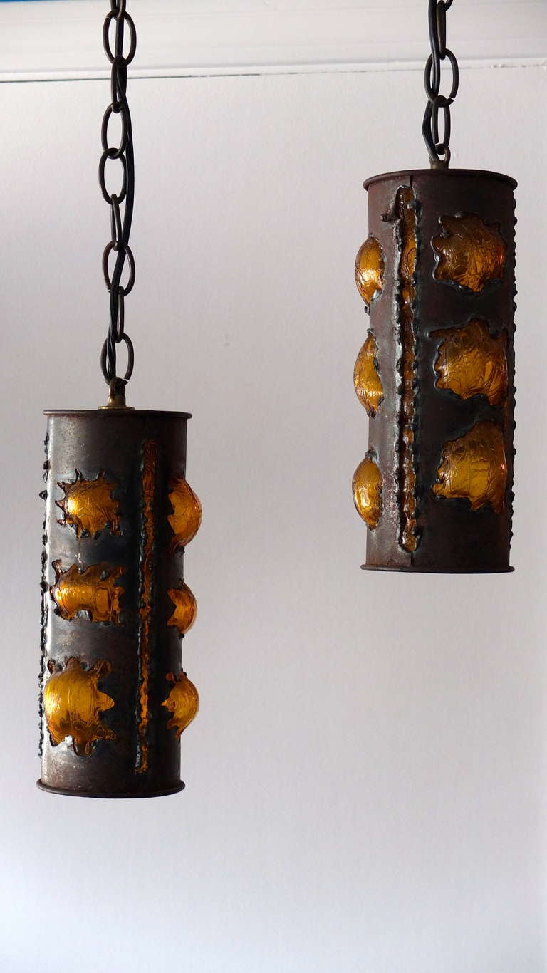 Handsome pair of torch cut steel sleeve-form cylinder pendants with irregular amber glass 'bubbles' protruding through asymmetric openings in the skin. The steel has a bronze finish.  The textured golden amber glass is a single tube-like form with