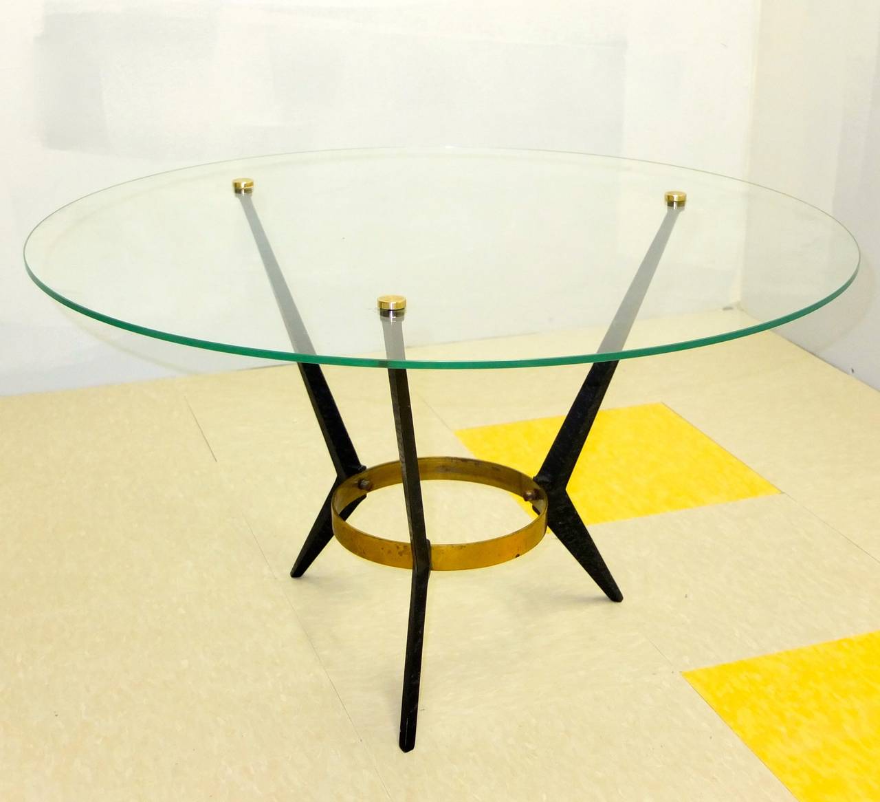 Early 1960s three leg cocktail table with round glass top by Angelo Ostuni, each leg made from cast aluminum finished in matte black in geometric angular tapered form around a round brass flat bar band. The round glass top is held in place by three