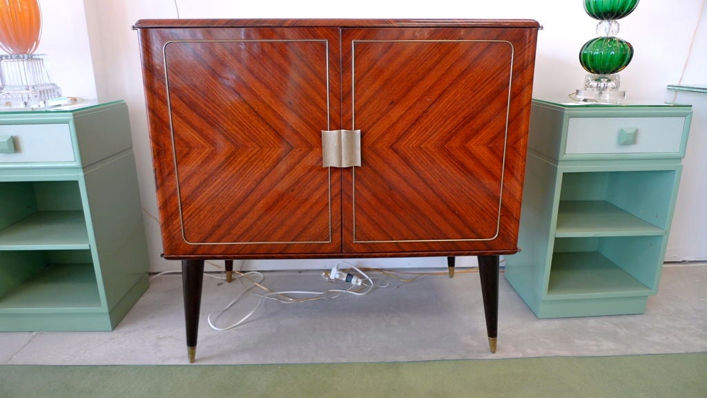 Beautiful gleaming Pallisander case with brass modernist hardware and brass inlay on the doors. Flared tapered legs with brass sabots.  Inside is gorgeous green glass and mirror back with single clear glass shelf with a recess curve to accommodate