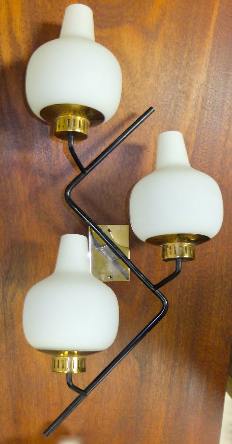 Signed near-pair of wall lamps by Stilnovo.  Similar to a design by G.C.M.E..  Black enameled brass zigzag structure with brass bobeche and mountings.  Gourd shaped opaline glass vessels.

We have a third lamp available separately with round white