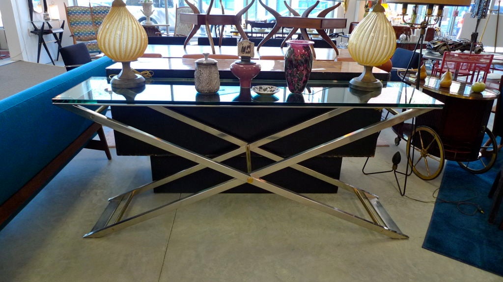 This X frame striking console has a polished glass top and a stainless steel base with brass accents.