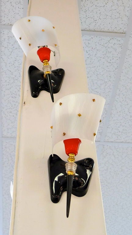 These are truly jolly with their amoeba form black bases, gold studded white ribbed scroll shades and both red and black torch stems held by brass and Lucite mounts. French glam deco! I would love to see them on red lacquered walls.<br />
We have