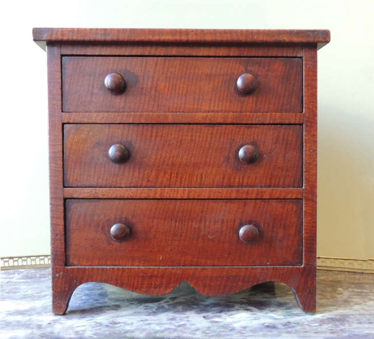 This unique piece is made was made as a sample for a salesman to be able to show his products. This miniature chest is made of Tiger Maple and has three drawers with six identical knobs. The bottom of this piece features a simplistic curved skirt.