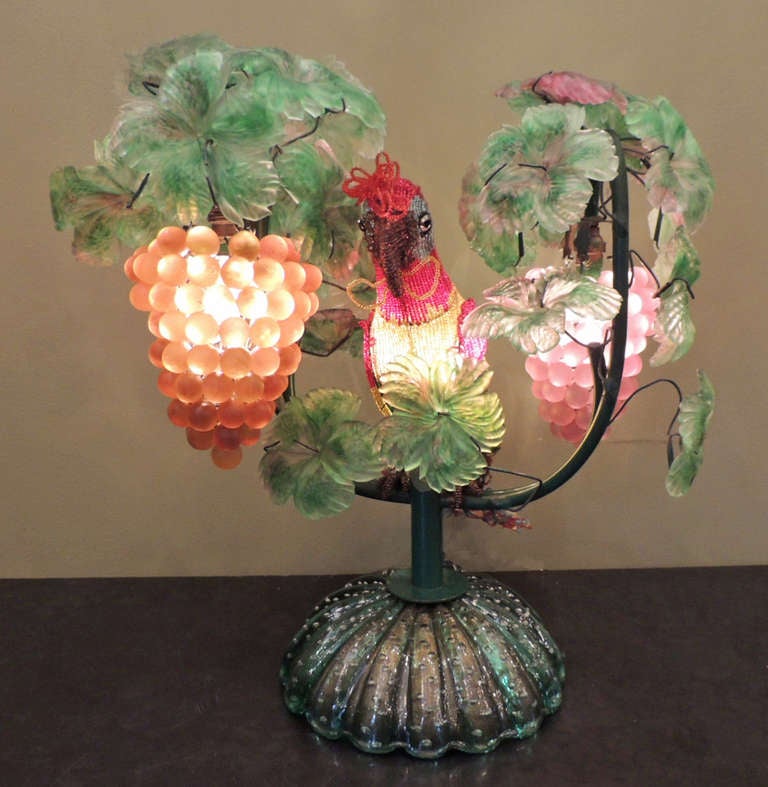 This colorful lamp was made on the Italian island of Murano, which is famous for its handmade glass items. There are two opaque grape bundles that are surrounded by leaves, and the central detail of this lamp is a colorful glass beaded parrot. The