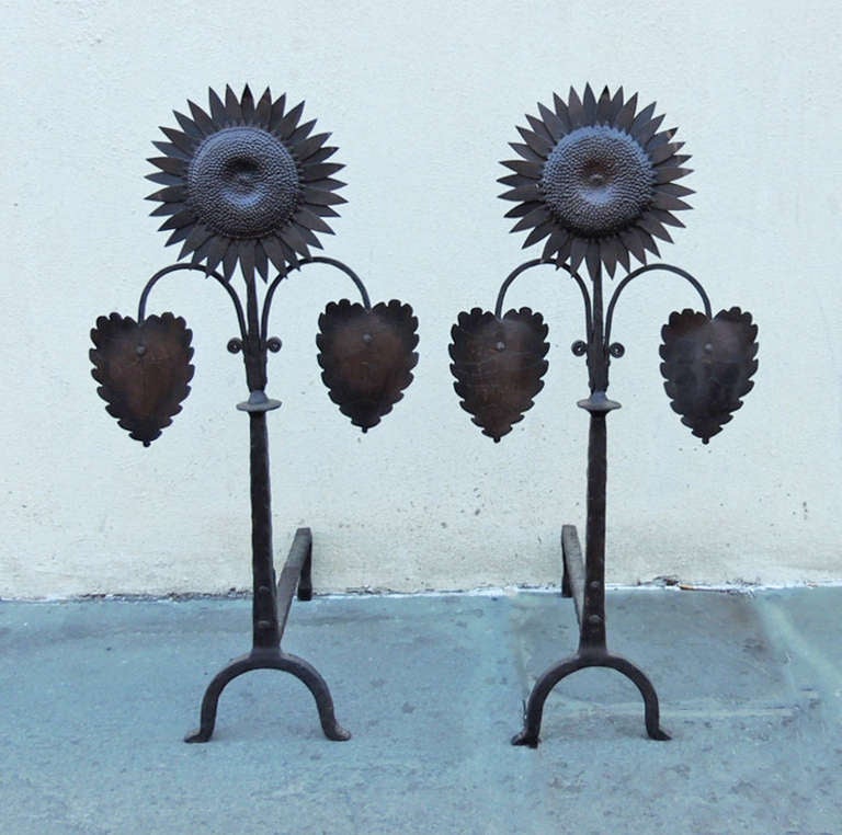 These andirons were made in the United States during the Arts and Crafts period of the early 1900s. They feature a detailed sunflower motif at the top and downward-pointing leaves.