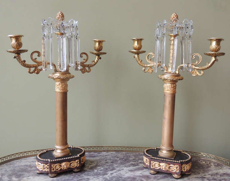 This pair of candelabras were made in France during the first half of the 19th century. The top of the pieces feature scrolling details on the arms and bobeches. Long crystal prisms hang from a finial at the top of the candelabras. The column-like