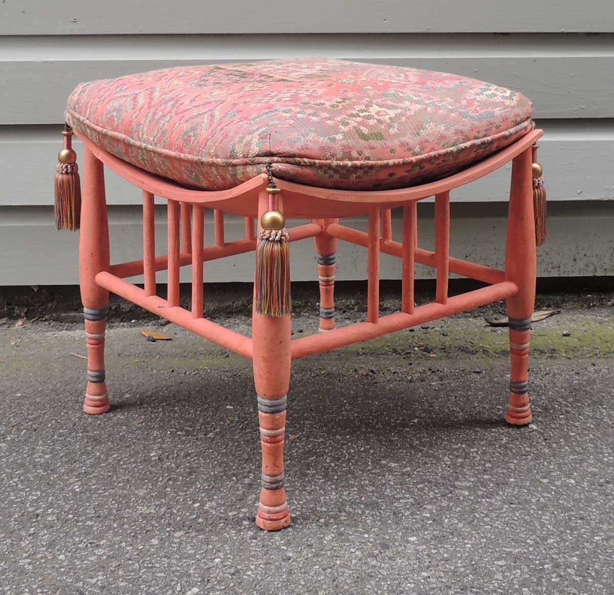 This unique stool was made in the first half of the 20th century and reflects the Egyptian style. This piece is painted a salmon pink with blue and white stripes on the legs. The cushion features similar colors with multi-color tassels at each