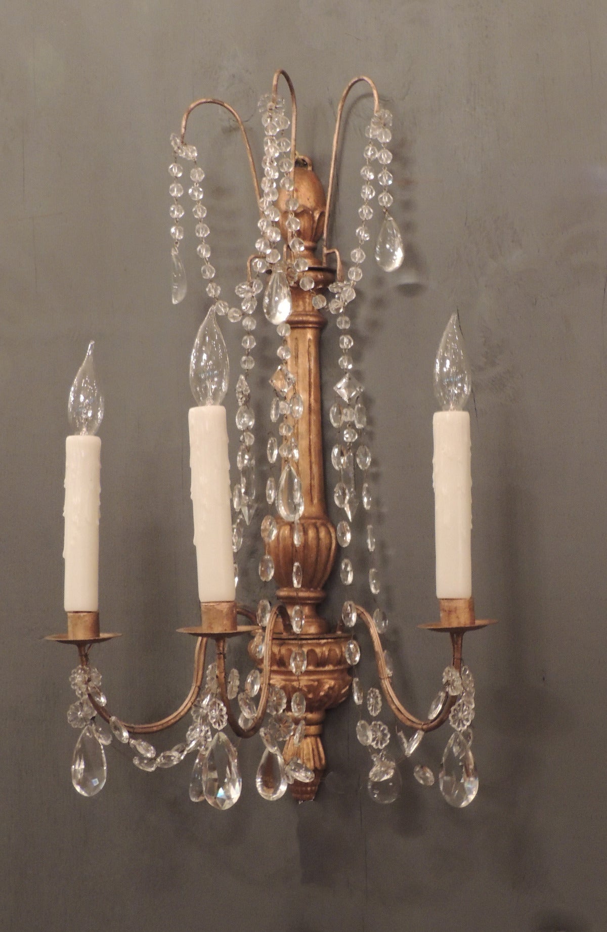 This pair of Genoese sconces were made Genoa, in Southern Italy, in the early-19th century. They feature a gilt carved stem with three candle arms, crystal swags and prisms, and an abstract corn finial. They have been rewired with new porcelain