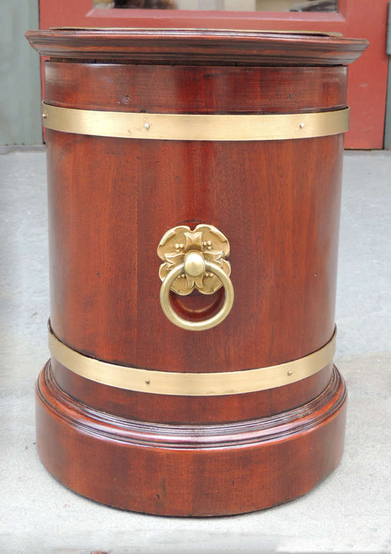 This wine cooler was made in England during the 19th century and is made of mahogany with double brass rims and handles. There is also a removable brass lining. The handle is in the shape of a flower with a central ring. There are two handles on the