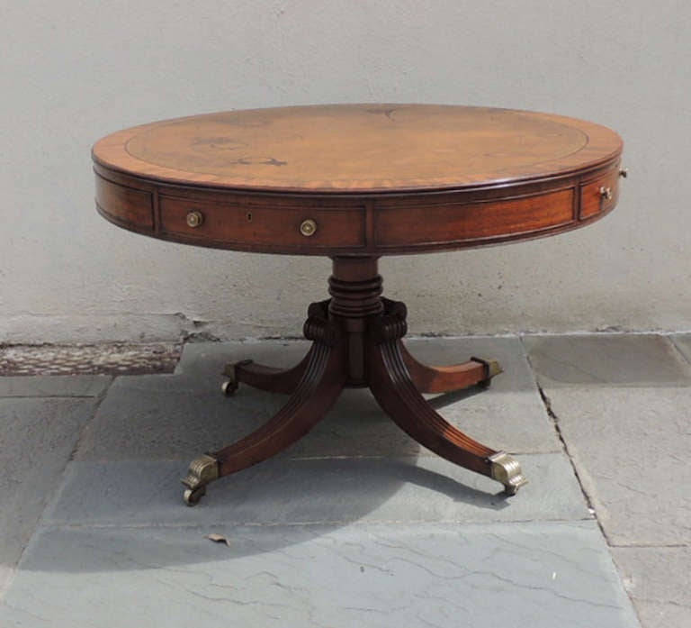 The base of this table is made of solid mahogany, while the top is constructed of mahogany and pine veneer with a brown leather writing surface. The leather top features a floral gold trim and has some wear due to age and use. The circular body of