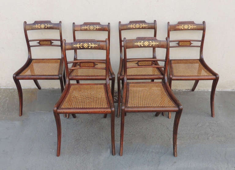 This set of six dining chairs is made of Rosewood with beautiful brass inlay. The ears are slightly more raised than the splat that features two horizontal panels. The top panel has carved details that border the brass inlay middle section.  The