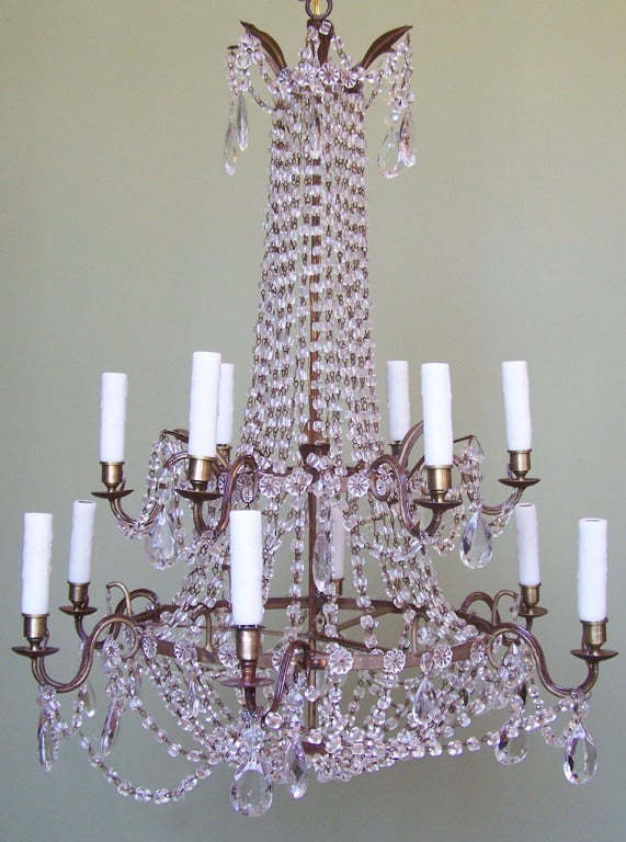 A late-18th century Italian empire chandelier featuring two tiers supporting twelve arms. Fine chains of crystal swags hang from the candle arms, bobeches, and decorative metal leaves. Larger crystal prisms provide extra sparkle to this piece.