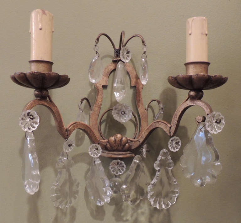 This pair of sconces were created in the early 20th century and are made of gilt and iron. The pieces have two candle arms hung with crystal prisms and rosettes.