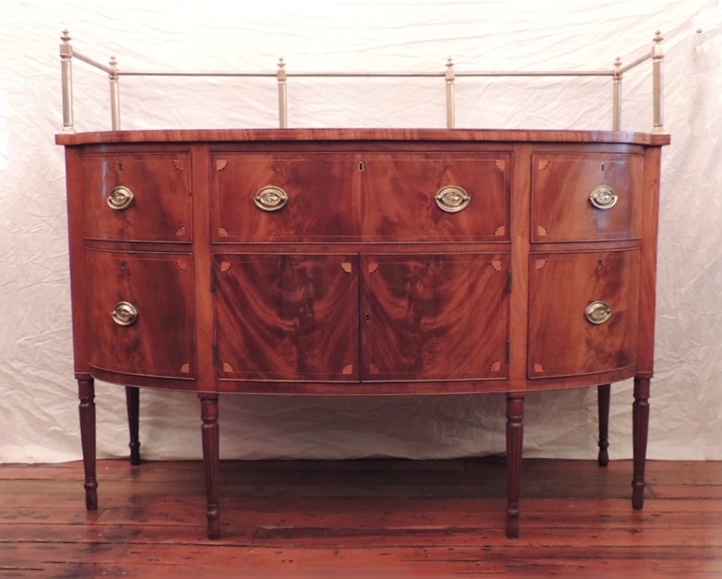 This Sheraton sideboard was made in America  circa 1800, and features a brass gallery, five drawers, and a cabinet.  The piece features lion pulls and reeded Sheraton legs. 