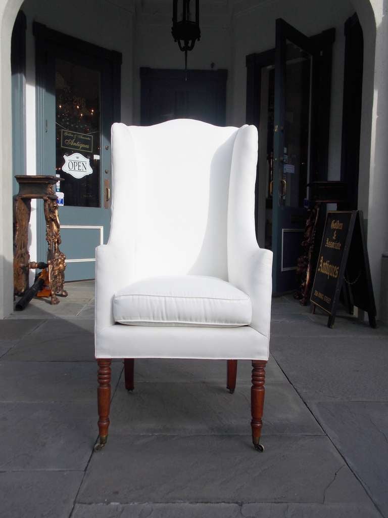American mahogany Sheraton wing back coachman's chair with serpentine back, flanking scrolled arms, and terminating on turned bulbous legs with the original brass casters. Chair is upholstered in white muslin. Early 19th century.