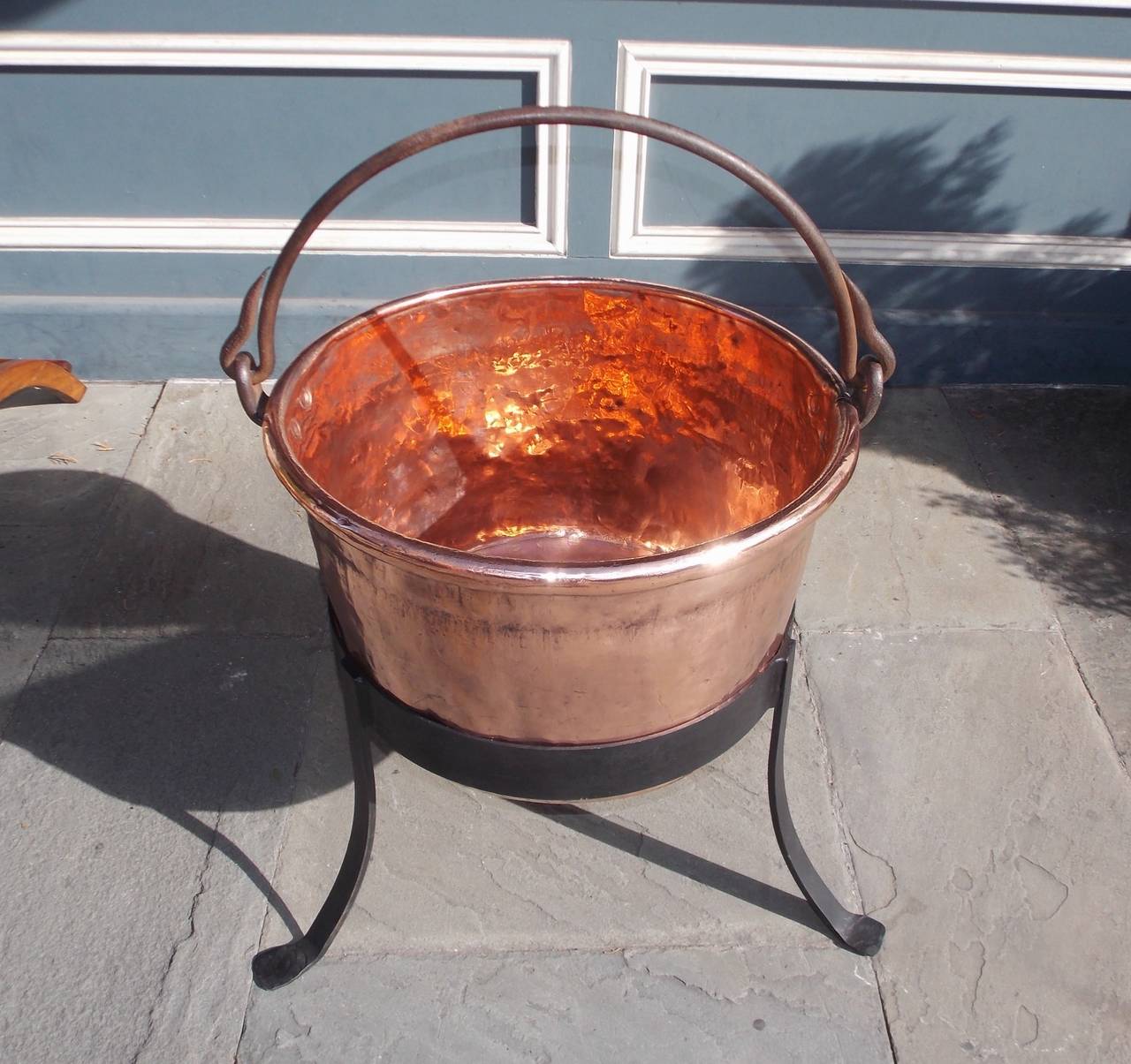 American copper and wrought iron plantation cauldron with original folding handle and supported by circular fitted three leg splayed stand. Late 18th Century