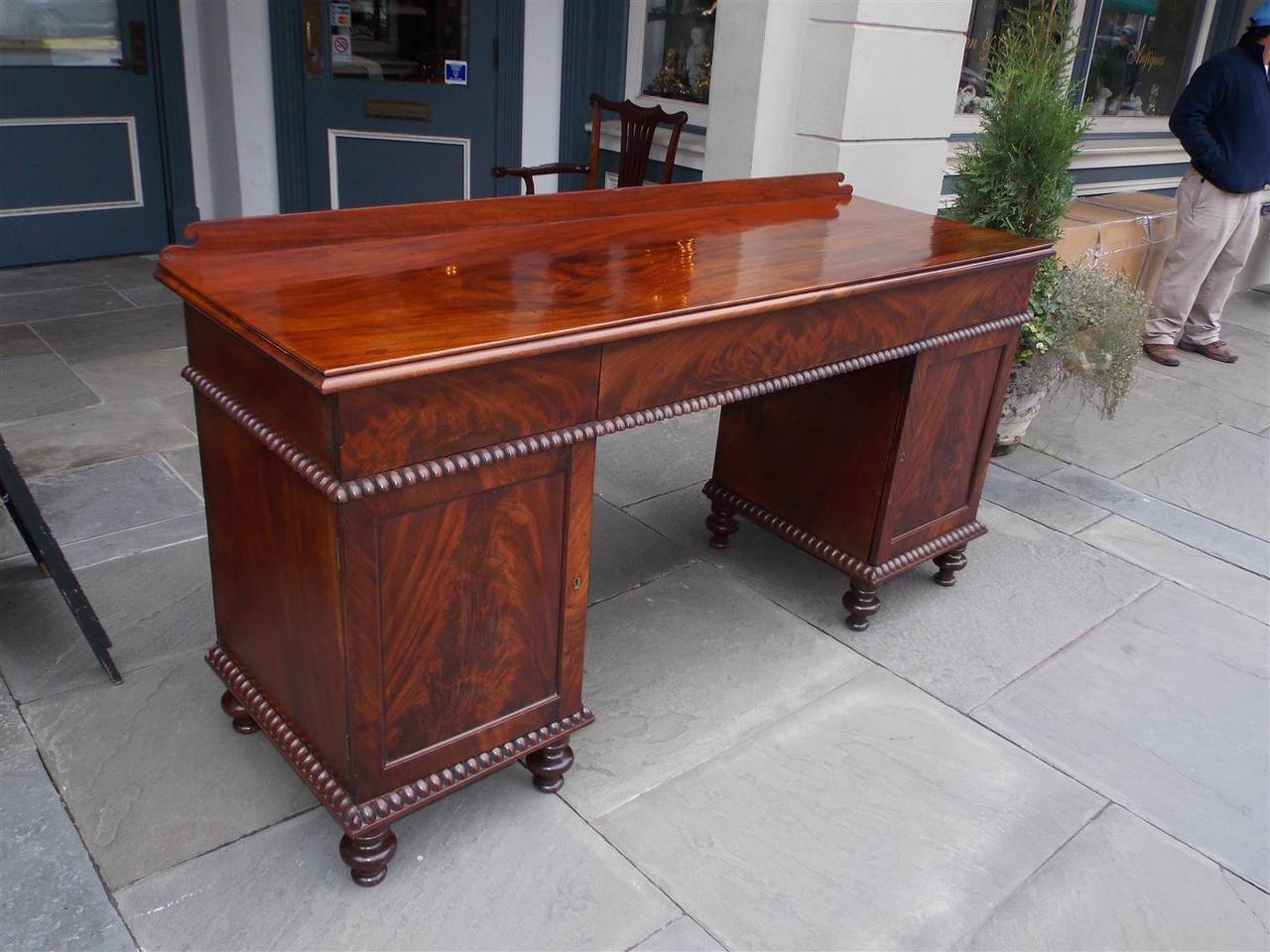 English Georgian crotch mahogany sideboard with carved scrolled back splash, three flush hidden upper drawers, lower flanking cabinets with interior shelving, gadrooned molded carved edges, and terminating on turned bulbous feet.
Early 19th century.