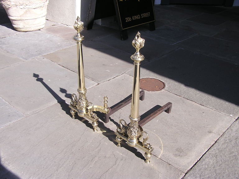 Pair of French brass andirons with flame finial top, tapered column with chased acanthus supported by flanking dolphin plinths on turned bulbous feet with matching log stops. Dealers please call for trade price.
