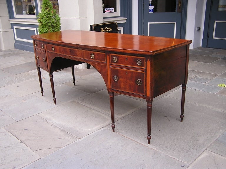 English Regency mahogany bow front sideboard with a one board top, central drawer with flanking cabinets, foliage brasses, satinwood inlaid panels with string inlays, and resting on the original turned bulbous reeded legs. Late 18th Century