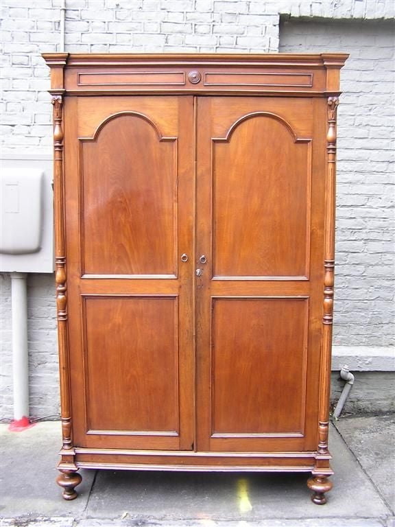 American Walnut hinged Armoire with a carved molded cornice, arched paneled doors, flanking acanthus carved bulbous columns, interior removable shelving, and resting on the original graduated bun feet. Early 19th Century