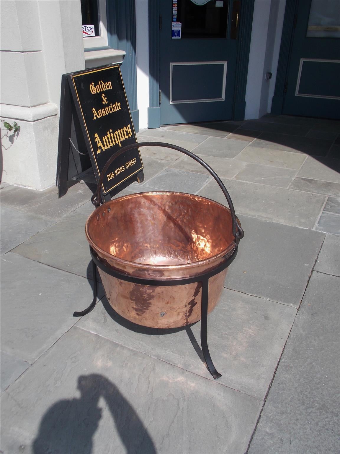 American copper and wrought iron plantation cauldron with original iron folding handle and supported by circular fitted three leg splayed stand. Late 18th Century