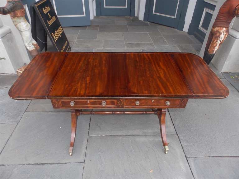 English mahogany and cross banded kings wood two drawer library table with satin wood string inlays, original brass pulls,  turned bulbous ringed ebonized cross stretcher, and resting on the original splayed legs with brass cup casters.  Early 19th