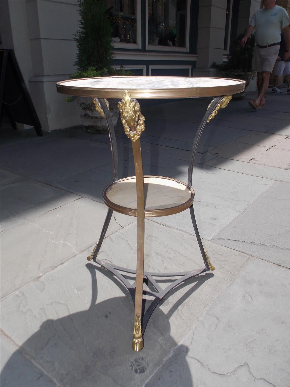 Italian marble and brass two-tiered bistro table with decorative gilt floral ram's head mounts and terminating on tripod legs with gilt hoof feet and wrought iron cross stretchers.  Early 19th Century.