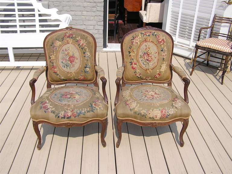 Pair of French Louis XVI walnut arm chairs with carved floral motif, original floral petit point, and terminating on stylized cabriole legs. Late 18th Century.