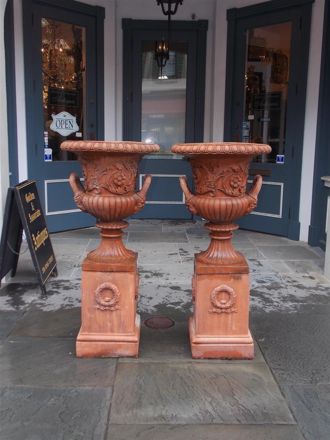 Pair of Italian terracotta urns with egg and dart molded edge, scrolled acanthus floral motif, molded side handles with figures of Bacchus and terminating on reeded and fluted circular bases resting on squared plinths with decorative laurel wreath