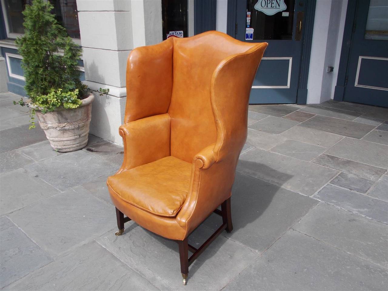 English mahogany caramel leather serpentine wing back armchair with front tapered and rear splayed legs, cross stretchers, and original brass cup casters. Late 18th Century.