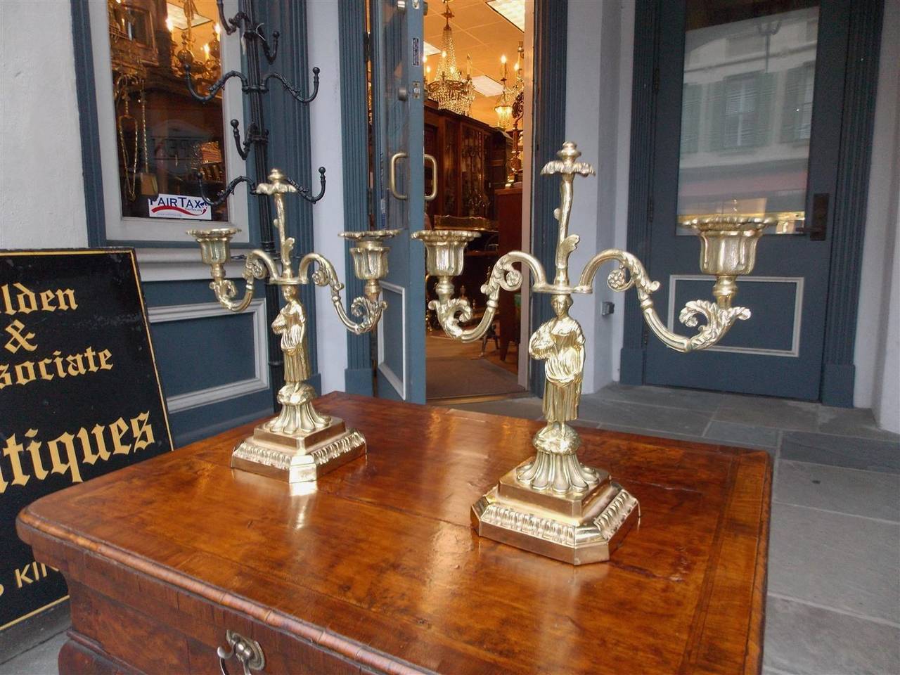 Pair of French brass two-arm figural candlesticks with ladies holding fruit baskets, scrolled floral arms, decorative floral medallions, original removable bobeches, and resting on squared plinths with lambs tongue motif. Candlesticks are candle