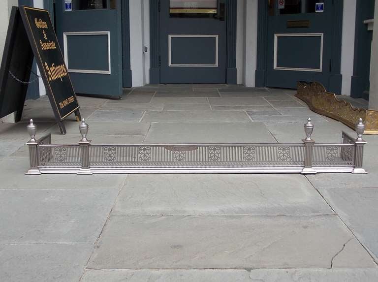 English polished steel fire place fender with flanking urn finials, engraved pierced foliage gallery with single row of beading, and resting on fluted step back plinths with a molded edge base. Last quarter 18th century