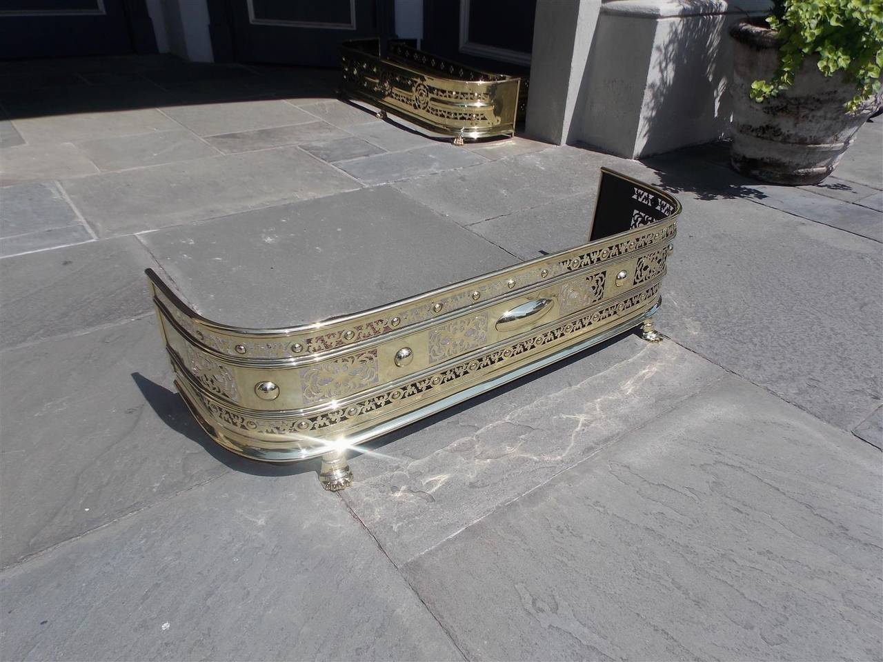 English Regency brass hand chased and bossed fire fender with circular medallions, floral motif, and terminating on stylized paw feet, Early 19th century.