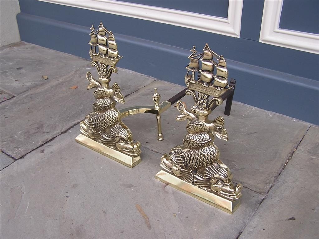 Pair of American brass andirons with perched flanking ships, intertwined dolphins, matching finial log stops , and resting on rectangular plinths. Mid -19th Century. 