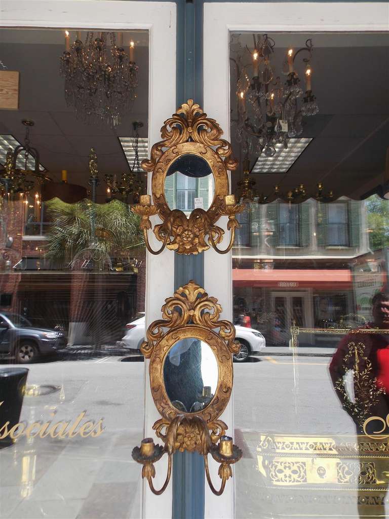 Pareja de apliques neoclásicos italianos girandole de madera dorada y gesso de dos brazos ovalados con espejo, con cresta de follaje en volutas, bordes interiores decorativos y brazos de vela con volutas de acanto, Principios del siglo XIX.  
Los