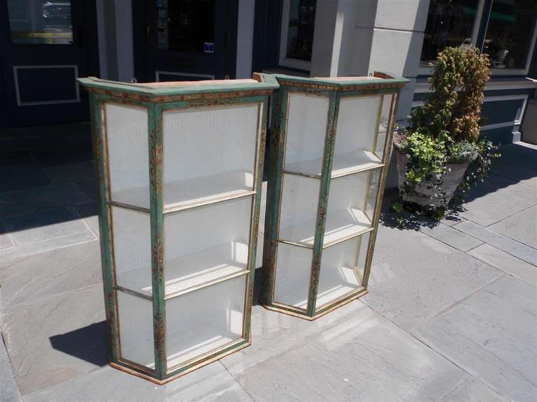 Pair of Italian painted and gilt hanging glass wall vitrines with interior upholstered shelving, foliage gilt stenciling, turn knob locking mechanisms, and retains the original wood backing.  Mid 19th Century