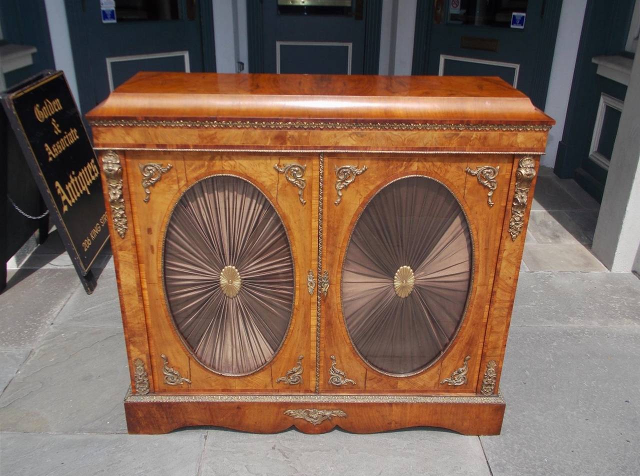 Pair of English burl walnut credenzas with floral ormolu mounts, flanking doors with original oval glass, silk curtains with centered ormolu medallions, and terminating on a rectangular serpentine molded edge base, Early 19th century.