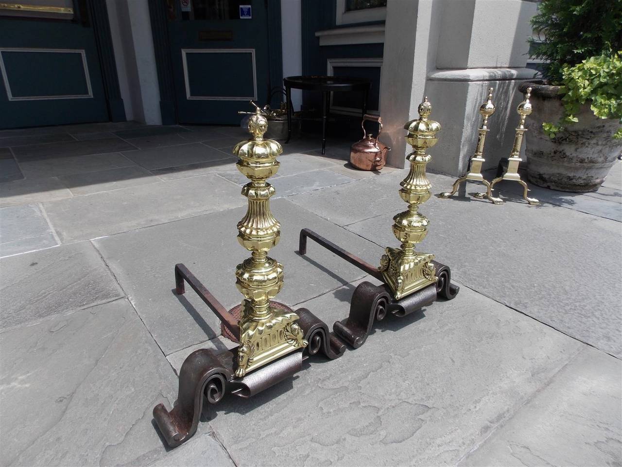 Pair of English brass and wrought iron andirons with hand chased faces on a fluted finial top, bulbous fluted centered column, triangular plinths with chased faces and lions paw motif and terminating on scrolled wrought iron legs, Mid-18th century.