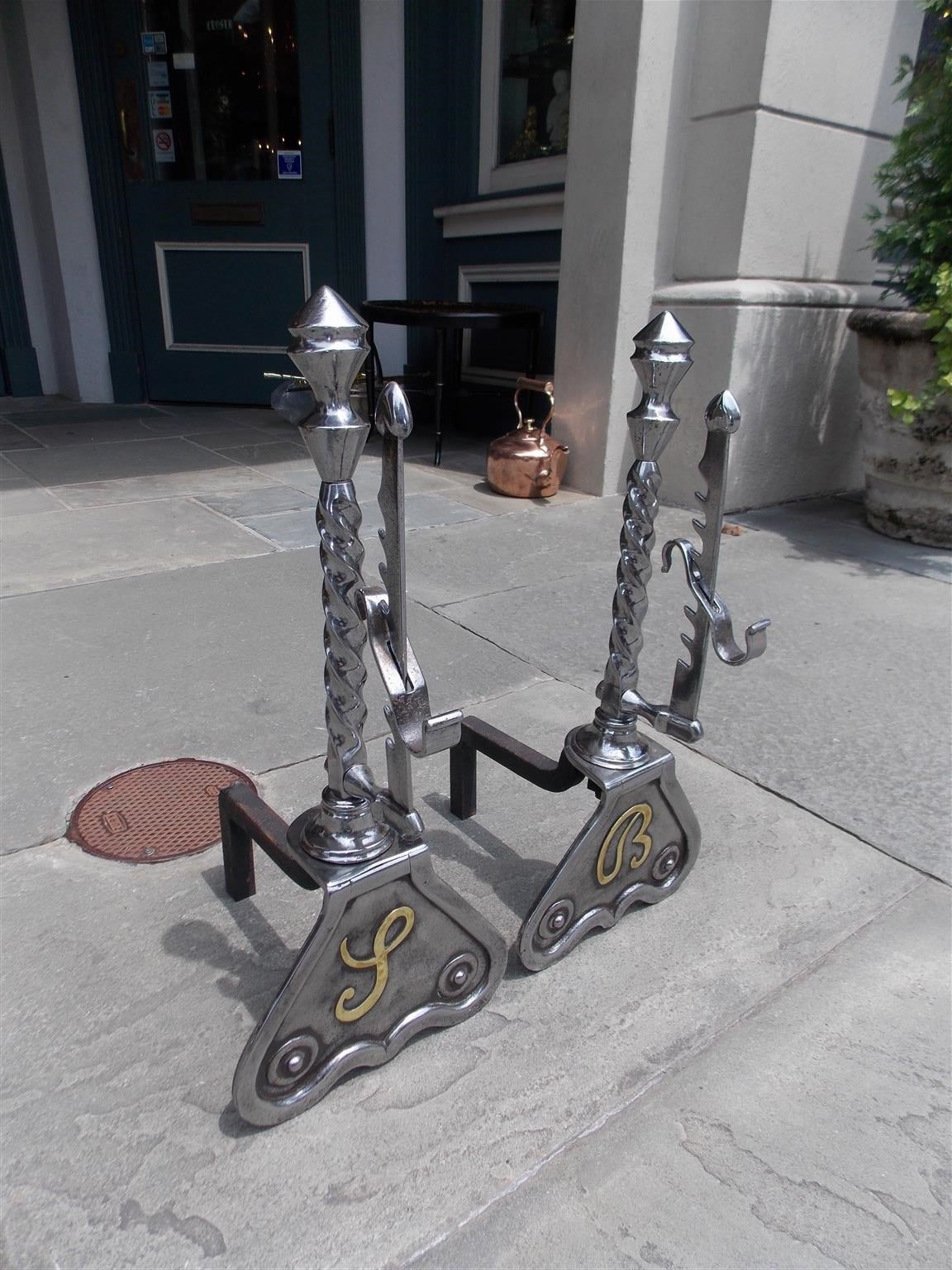 Pair of English polished steel and nickel silver andirons with a urn top finial, twisted centered column, adjustable spit hooks,  and terminating on scrolled brass monogrammed plinths.  Mid 19th Century