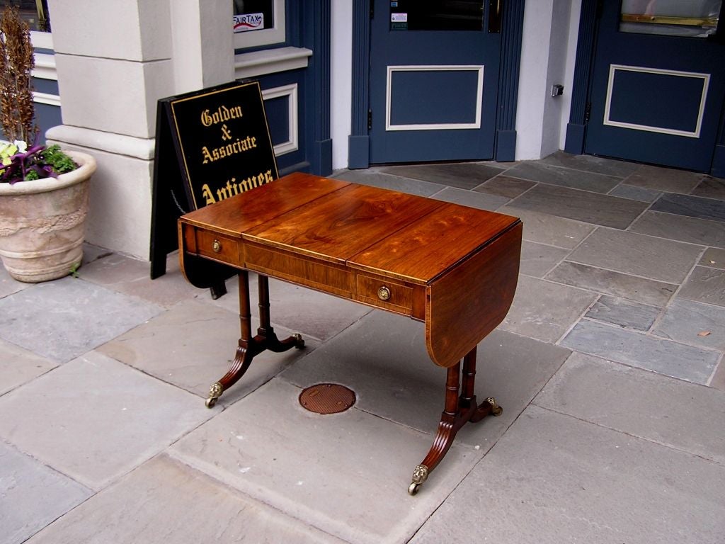 English Kings wood drop leaf two drawer library / games table with fitted leather backgammon game board,  flip top satinwood inlaid checker board , double  turned reeded ringed legs, and terminating on the original lions head brass casters. Late