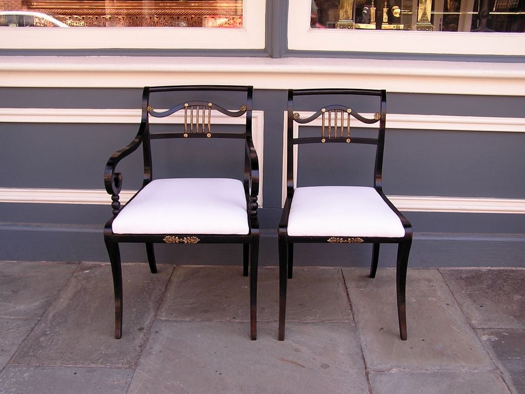 Set of Eight English Regency painted and silver gilt dining room chairs with scrolled arms, original ormolu mounts, and resting on saber legs. Late 18th Century. Dining room chairs have the original seat frames and are upholstered in white muslin. 