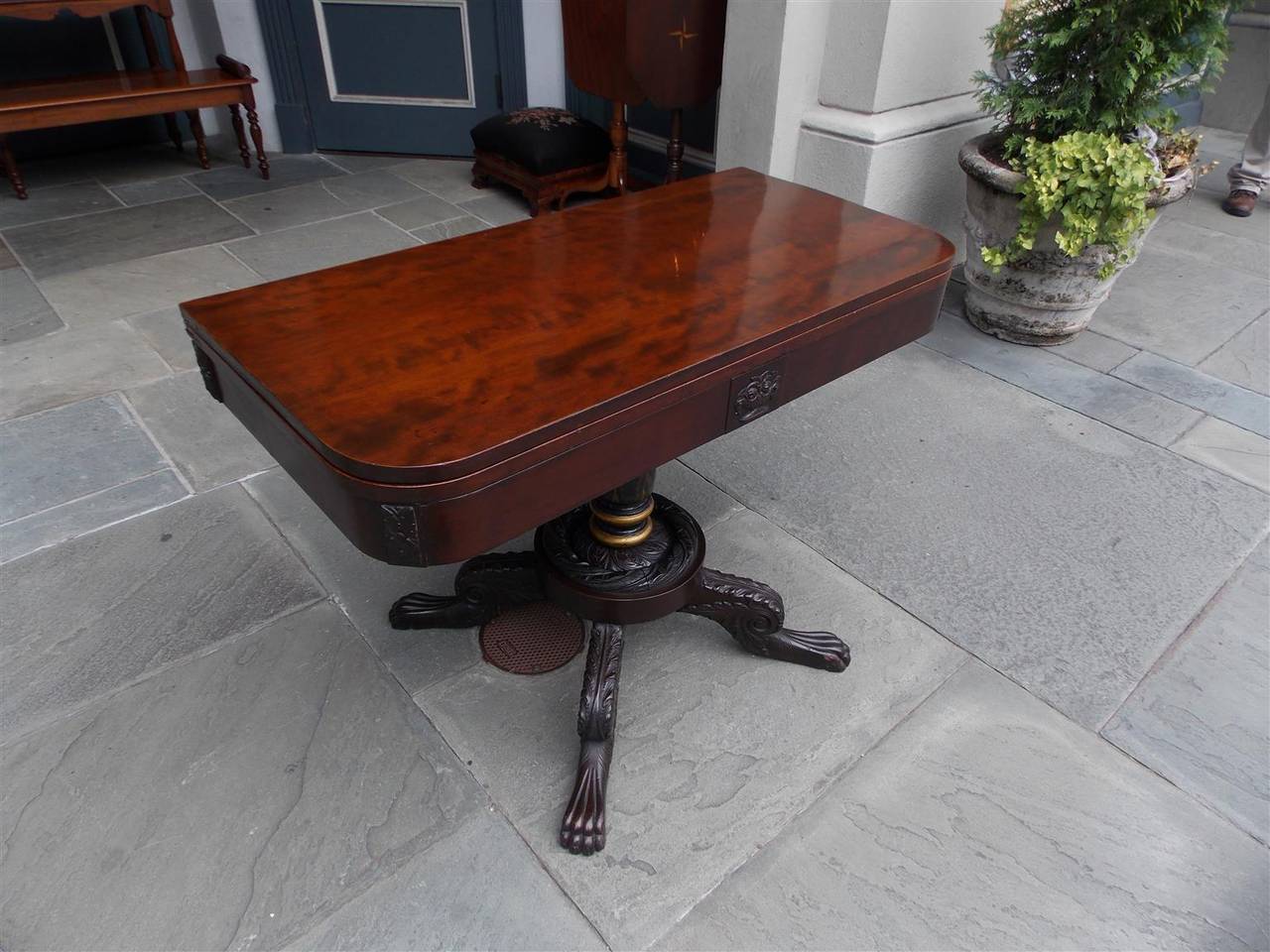American Federal mahogany and cherry hinged game table with a one board top, central carved floral basket, stenciled gilt turned ringed center column, and resting on scrolled acanthus carved legs with lions paw feet, Philadelphia, Early 19th Century.