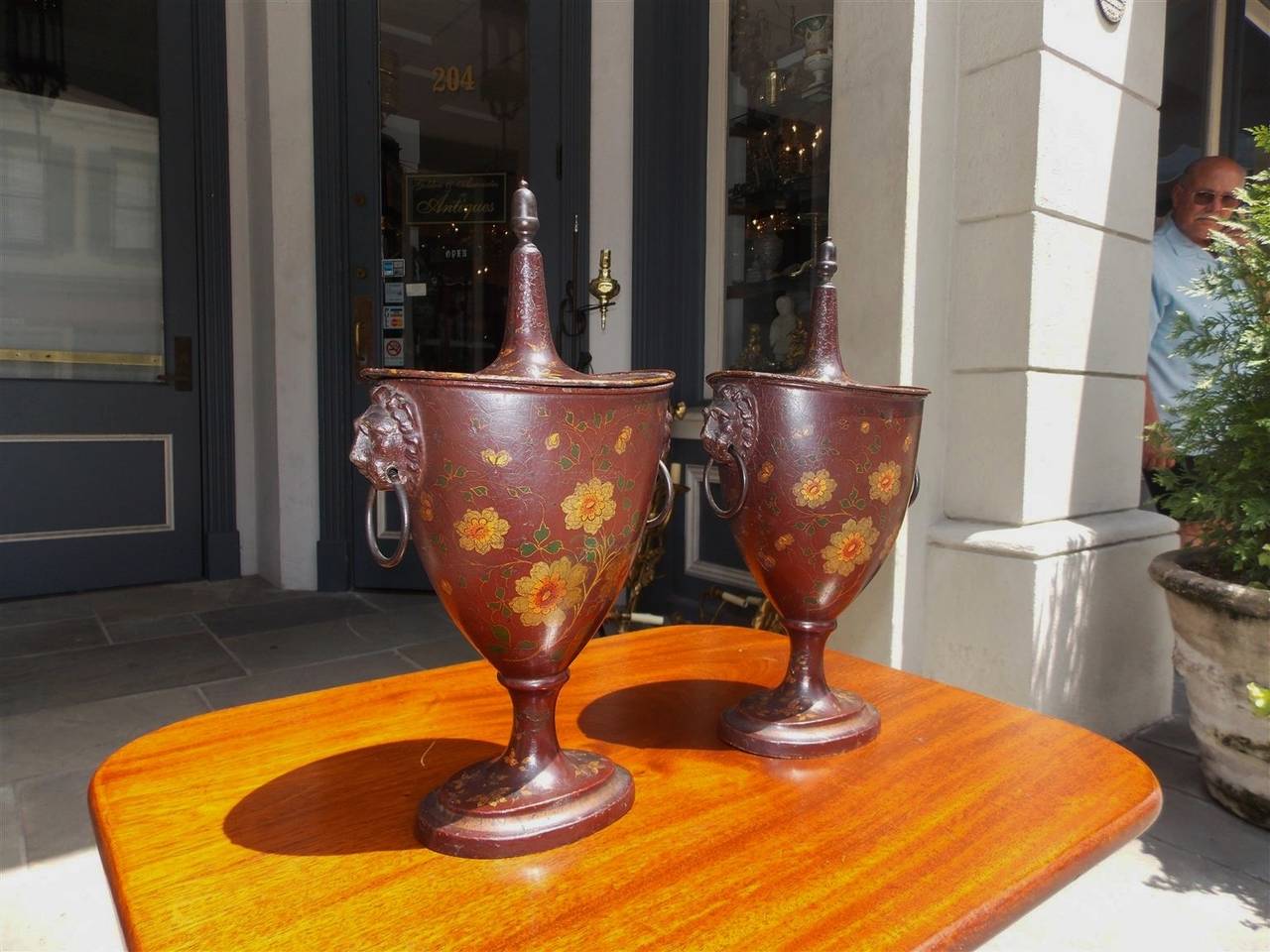 Pair of English Regency tole chestnut urns with hand-painted floral decorative motif, removable lids with acorn finials, original lions head side handles with rings and terminating on a circular molded edge plinths, Early 19th century.