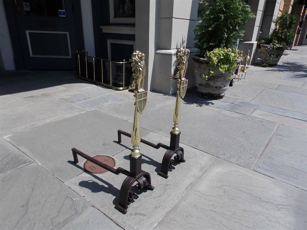 English brass and wrought iron shield perched lion andirons with turned bulbous brass columns, rectangular iron plinths, and terminating on chased scrolled legs, Early 19th century.