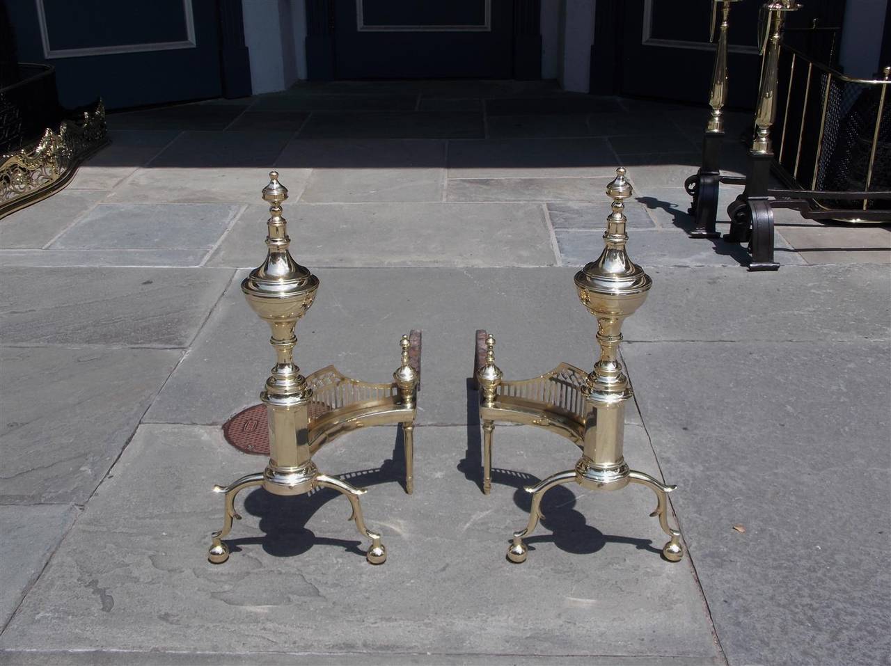 Pair of American brass urn finial andirons with turned bulbous faceted plinths, matching urn log stops, flanking pierced galleries, double spur legs, and terminating on ball feet.  NY, R. Wittingham, Early 19th Century.