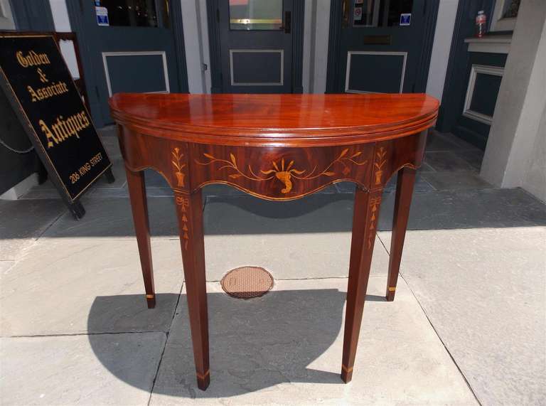 American mahogany flip top demilune game table with serpentine carved skirt, satinwood floral, ribbon, and graduated bell flower inlay terminating on tapered cuffed legs table has one board tops. Late 18th century. Table is 35.5