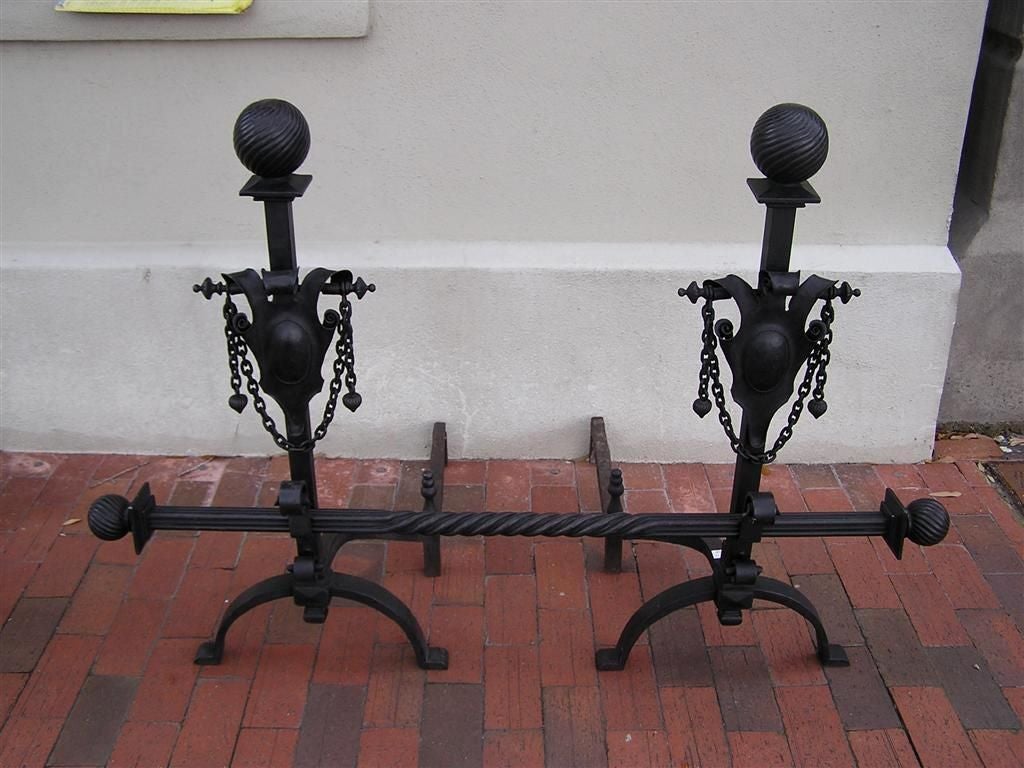 Pair of American wrought iron spiral ball finial andirons with flanking medallion shields supporting hanging finial chains, scrolled spit hooks, decorative centered spiral crossbar, curvature supports with matching logs, and resting on scrolled legs