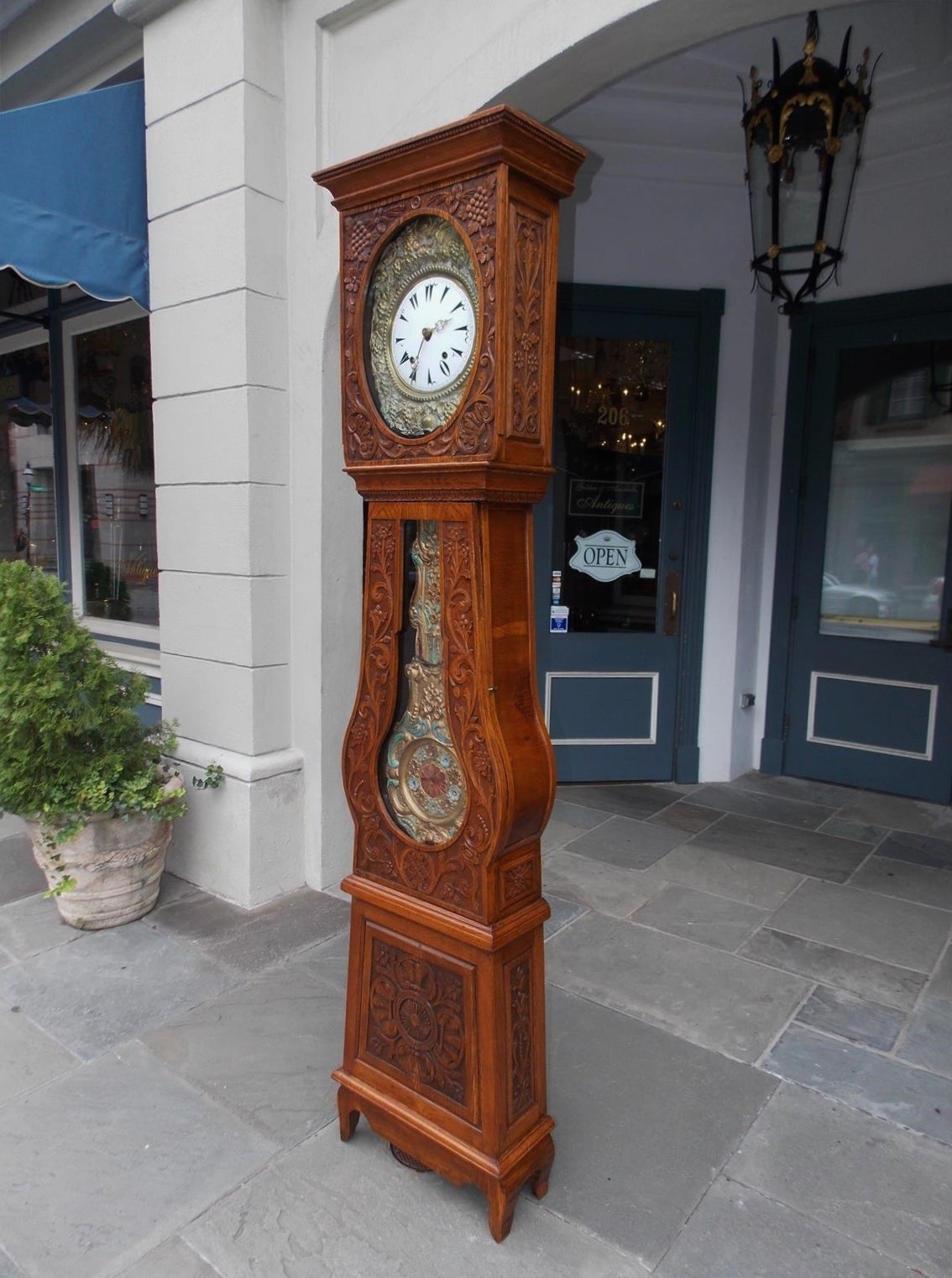 French Morbier oak tall case clock with hand-carved grape vine, floral and shell motif. Clock has a hammered brass and enamel painted dial under glass, brass and hand-painted pendulum under glass and terminates on a squared plinth with cone feet,