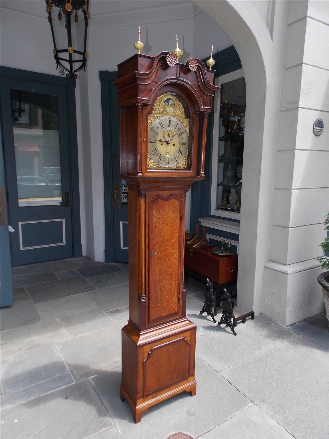 English Chestnut and Mahogany tall case clock with broken arch pediment, floral rosettes, original brass urn finials, dolphin and figural ormolu face, original arched hood with glass door, cross banded trunk and terminating on rectangular molded